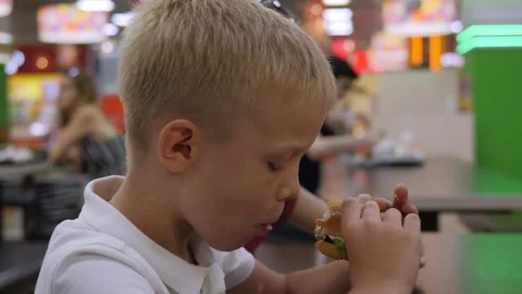 Close-up of a boy eating a burger sitting at a table in a shopping center. Stock Footage 296270475