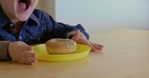 A close-up of a boy smelling a donut glazed with beige chocolate. The boy is at Video stock 347028704