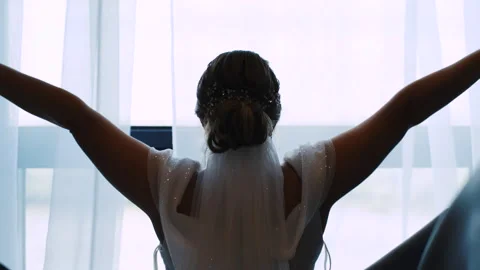 Close-up of bride in wedding dress opens huge curtains and looks out the window 動画素材 285436440