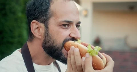 Close up a brunette guy with a beard bites a hot dog that he cooked on a Stock Footage 283832373