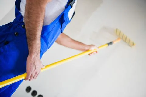 Close up, a builder in overalls paints a wall in white with a roller Stock Photos