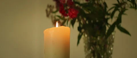 Close-up of a burning candle in front of red flowers in vase in a dimly lit room Stock Footage 316765365