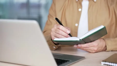 Close up of businessman's hands taking notes in notebook while using laptop  Stock Footage 295039008