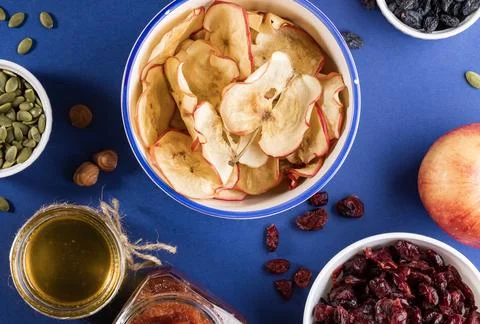 Close-up ceramic cup with apple chips surrounded by healthy snacks on blue .. Stock Photos