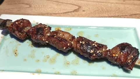 Close-up of Charred Grilled Meat Skewer on Ceramic Plate Over Wooden Tabletop Vidéo 344853155