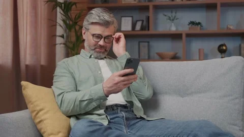 Close up of cheerful middle aged man with grey hair sitting on the sofa using Vídeos de archivo 220201340