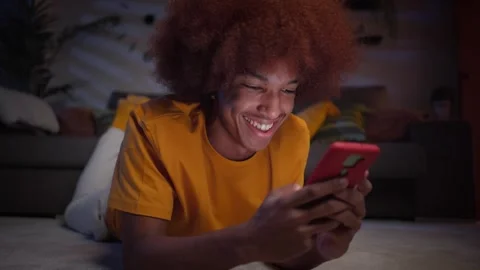 Close up cheerful young afro boy using smart phone lying on carpet. Smiling man Stock Footage 248866040