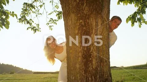 CLOSE UP Cheerful young couple peeking from behind a tree smiling for the camera Stock Photos