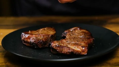 Close-up, The chef adds pepper to two juicy pieces of steak on a black plate Video stock 188310100