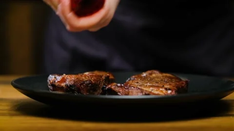 Close-up, The chef adds pepper to two juicy pieces of steak on a black plate Stock Footage 188567449