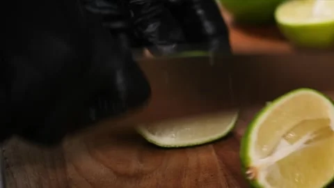 Close-up of a chef in black gloves slicing a fresh green lime on a wooden cuttin Stock Footage 331635499