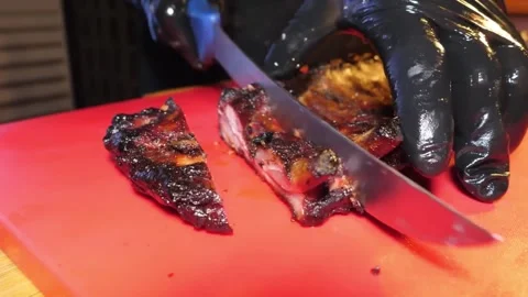 Close-up of Chef in Black Gloves Slicing Juicy Grilled BBQ Spare Ribs on a Red C Stock Footage 338212813