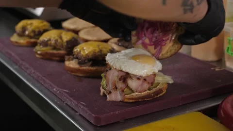 A close-up of a chef in gloves putting buns with sauce on burgers with fried egg Stock Footage 287587513