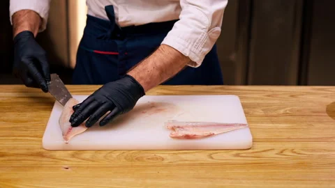 Close up a chef in a white jacket in a professional kitchen cuts fish with knife Stock Footage 273638610