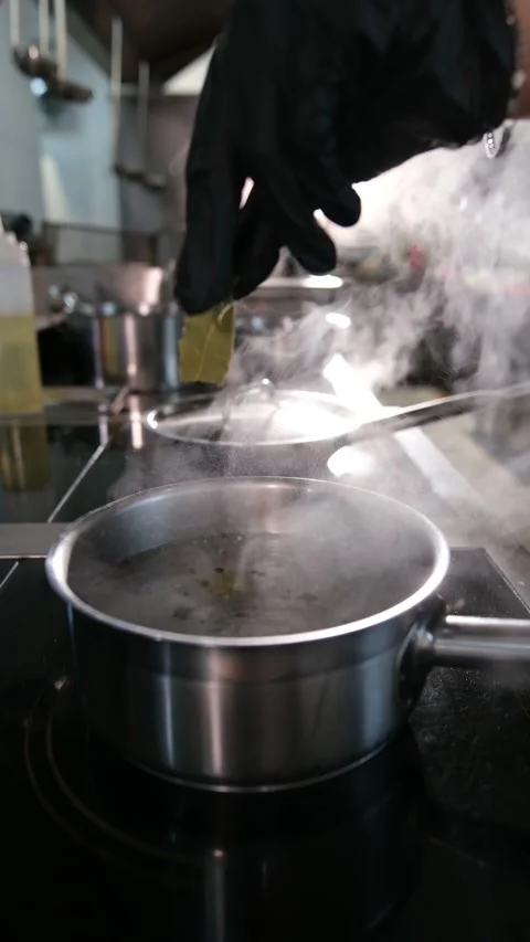 Close up of a chefs hand in a black glove adds bay leaves to a stainless pot of Stock Footage 318316982