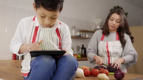 Close up of child in apron in kitchen playing with tablet while mother is coo Stock Footage 150664480