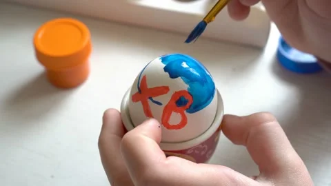 Close-up, the child paints a white Easter egg with a brush and blue paint. The 스톡 동영상 103100481