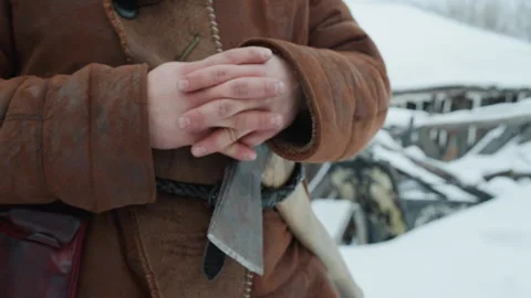 Close up of clasped hands under brown coat with axe beside in snowy ruins Video stock 347082144