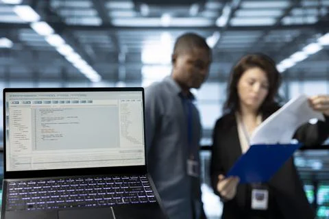 Close up of code on laptop screen next to server hub employees reading paperwork Stockfoto's