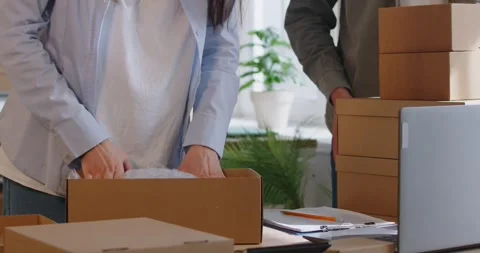 Close Up Of Colleagues Hands Packing Parcel Into Cardboard Box At Warehouse Stock Footage 280224136