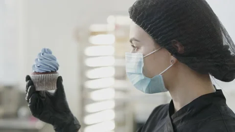 Close-up of concentrated young baker in Covid face mask examining baked muffin Stock Footage 150906920
