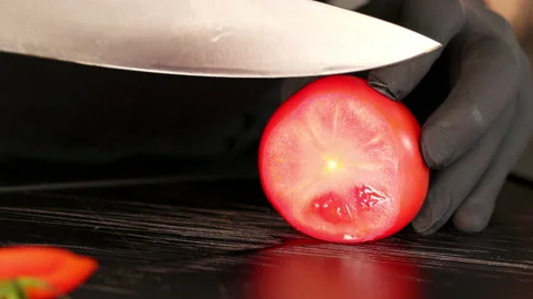 Close-up of a cook slicing a tomato. He uses a knife and cuts on a black board. Stock Footage 163046208