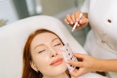 Close-up of cosmetologist using stencil to mark up woman face in preparation for Stock Photos