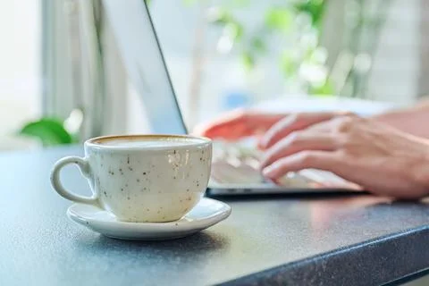 Close-up of cup with coffee on table, background is laptop, hands of man typing Stock Photos