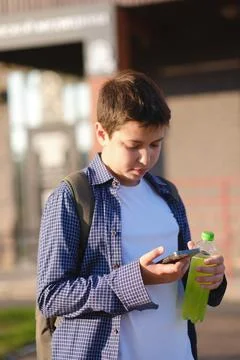 A close up of cute student boy with backpack holds a mobile phone and bottle of Stock Photos