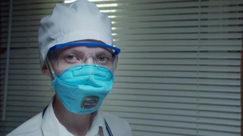 Close-up Doctor at a window with blinds in protective glasses and mask puts on Stock Footage 131620283