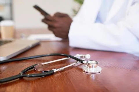 Close up of doctor's stethoscope while Black doctor uses cellphone in background Stock Photos