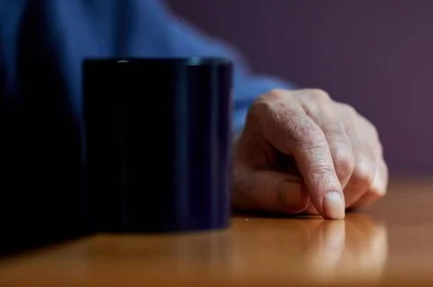 Close up of an elderly person's hand resting on a wooden table next to a dark Stock Photos