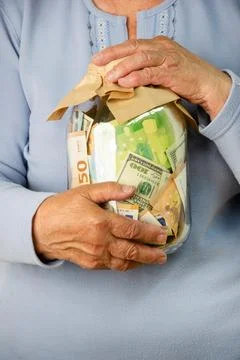 A close-up of an elderly womans hands holding a glass jar filled with euro and Stock Photos