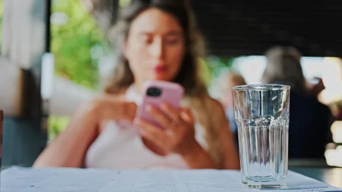 Close up of an empty glass on a table with a blurred view of a woman scrolling Stock Footage 314040663