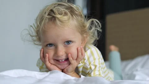 Close up of face of cute beautiful blue-eyed blonde baby child kid girl on bed Stock Footage 242401283
