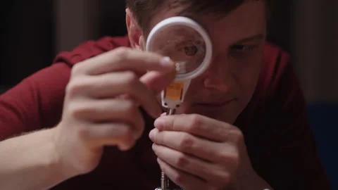 Close-up face of young man using magnifying glass to inspect small electronic Stock Footage 348084283