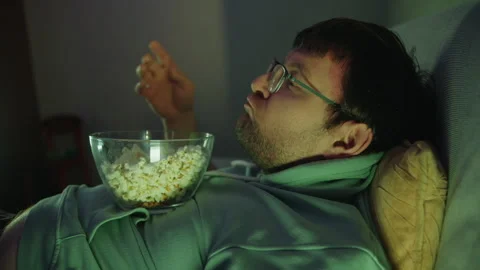 Close up of a fat man that takes popcorn and eats it while watching TV at night Stock Footage 314464244