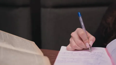 Close Up of Female Hand Writing in Notebook Beside Open Vintage Book Stock Footage 310698446
