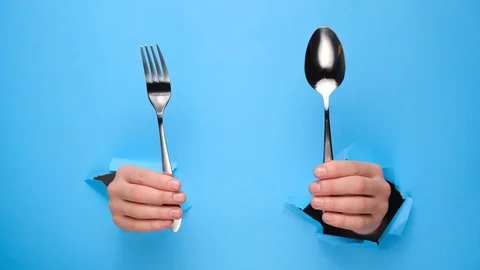 Close up of female hands holding spoon and fork through torn blue paper wall. Stock Footage 122171396