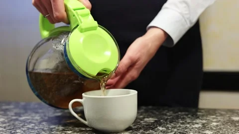 Close-up female hands pour tea from a glass teapot into a white mug on a black Stock Footage 273619595