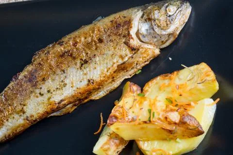 Close up of fresh fried trout with rustic potatoes on a black plate Stock Photos