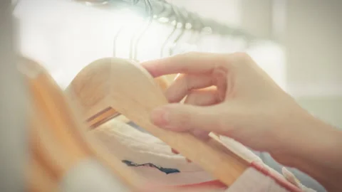 A close-up of a girls hands browsing clothes on wooden hangers on a gleaming Stock Footage 341490840