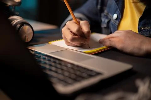 Close-up of girl's hands taking notes with pencil in notebook, laptop Fotos de archivo