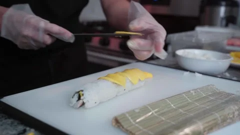 CLOSE UP OF GLOVED HANDS ADDING MANGO SLICES TO SUSHI ROLL ON CUTTING BOARD 스톡 동영상 336969985