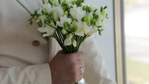 A close-up of the groom in a white jacket, holding a wedding bouquet of white Stock Footage 322183763