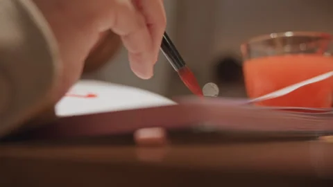 Close up of hand applying red paint with dip brush onto textured paper on wooden Stock Footage 345069994