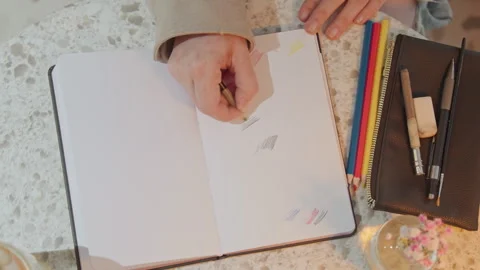 Close up hand drawing lines and shapes in open notebook surrounded by colored Stock Footage 345089809