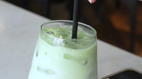 A close-up of a hand holding a black straw in a glass of iced green matcha latte Stock Footage 310218328