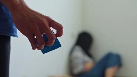 Close up of hand holding blue condom packet while woman sits distressed in Stock Footage 333514132