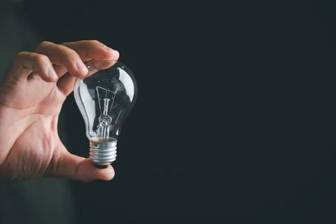 Close-up of a hand holding a clear light bulb against a dark background.  T.. Foto stock